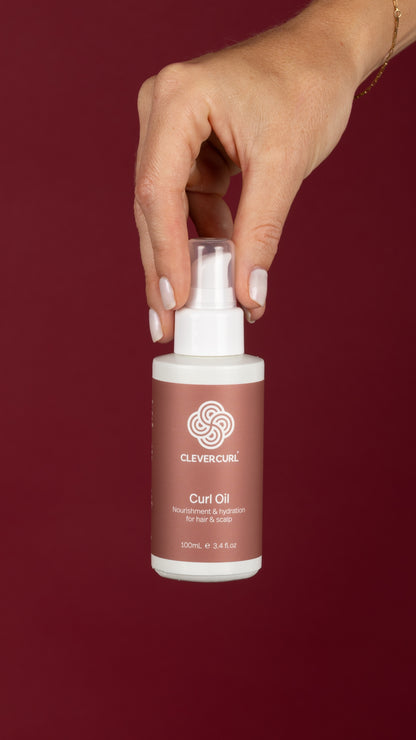 Hand holding a bottle of Clever Curl Curl Oil against a red background